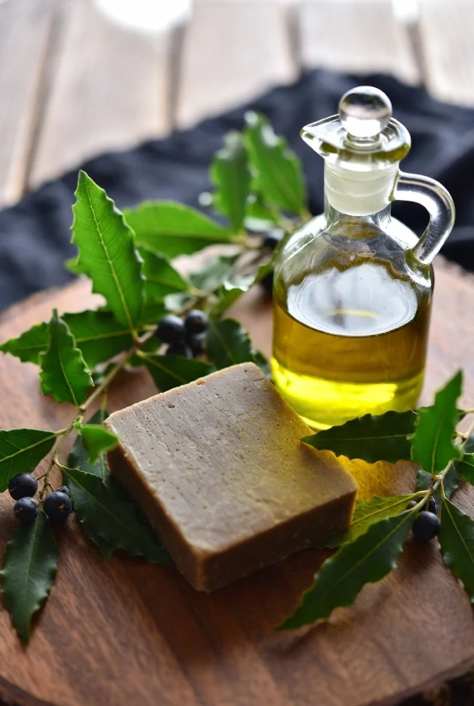 Close-up of authentic Aleppo laurel soap bar next to a glass bottle of olive oil, fresh laurel leaves and laurel berries on a wooden surface, natural daylight, very realistic food/product photography style.